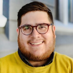 headshot of Andrés Serrano Chicago therapist in glasses, with brown hair and facial hair smiling at camera wearing a yellow shirt