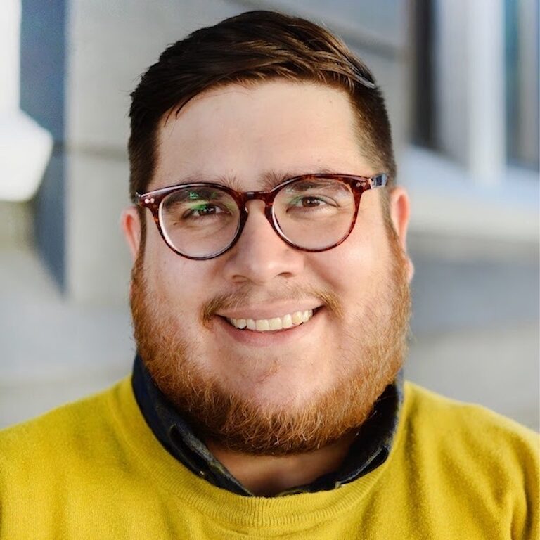 headshot of person in glasses, with brown hair and facial hair smiling at camera wearing a yellow shirt