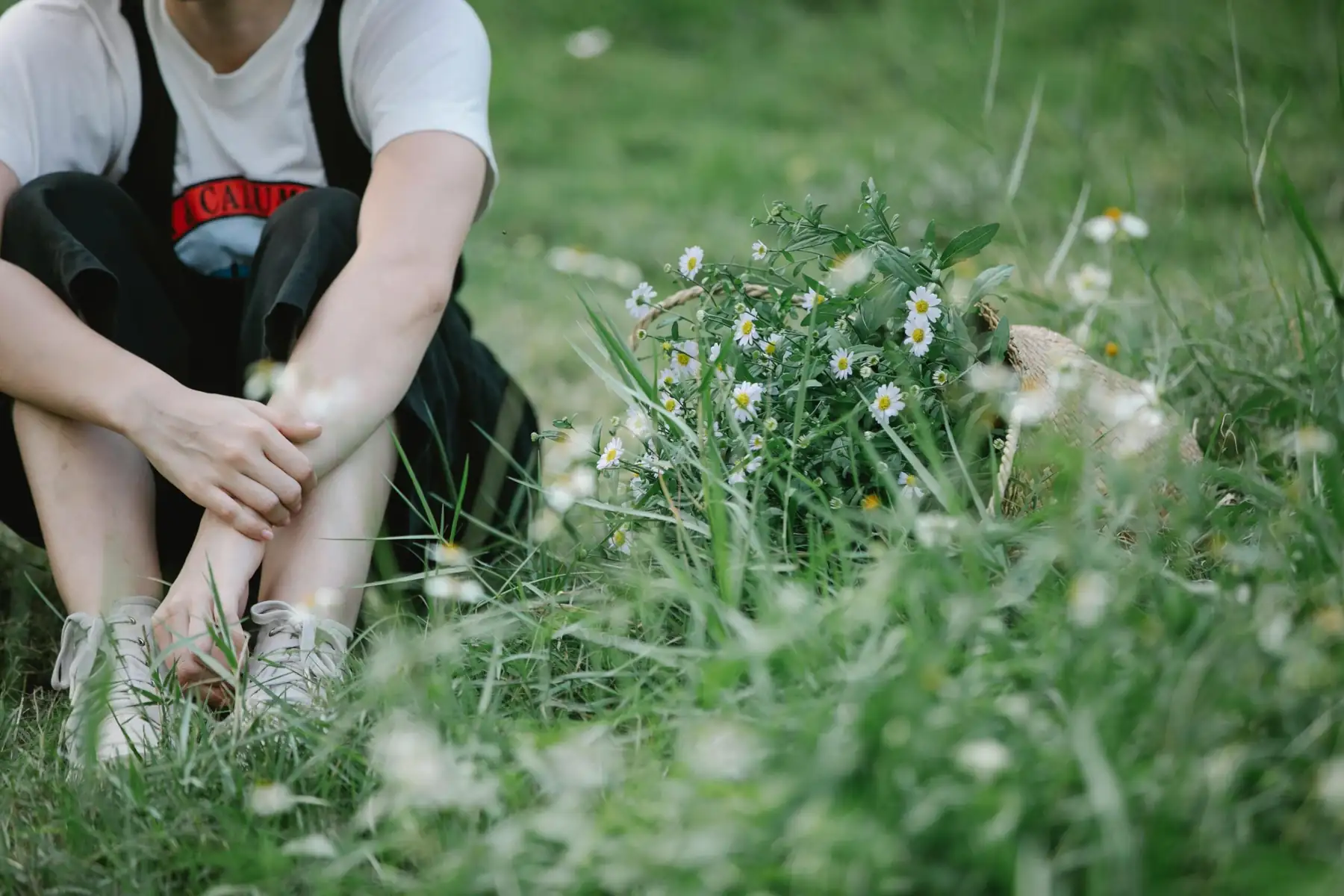 a person sits in the grass with their knees pulled to their chest, their face is not visible