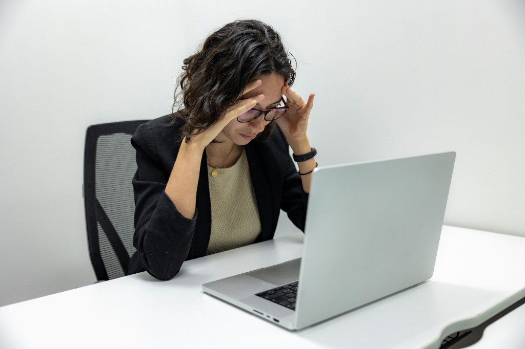 Woman in glasses at a laptop desk holding her temples, looking stressed or overwhelmed