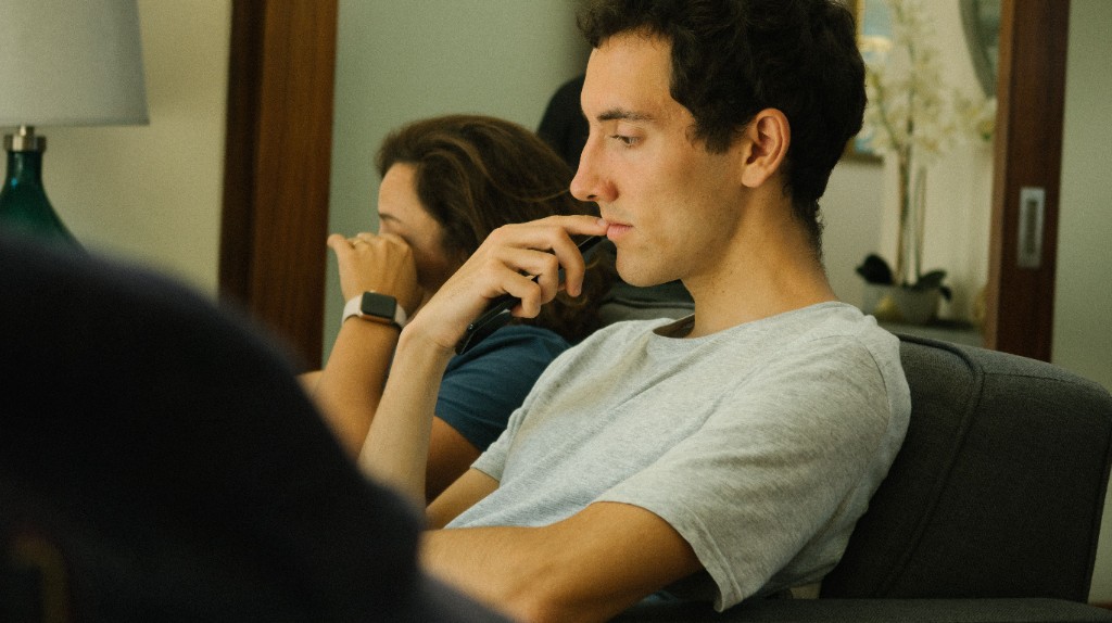 Young man sitting in a chair holding a phone to his lips thoughtfully while someone sits blurred in the background