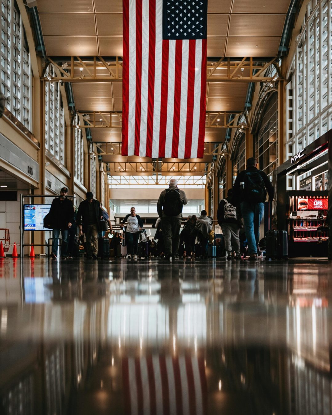 people walking under a huge american flag hung horizontally in an airport hallway